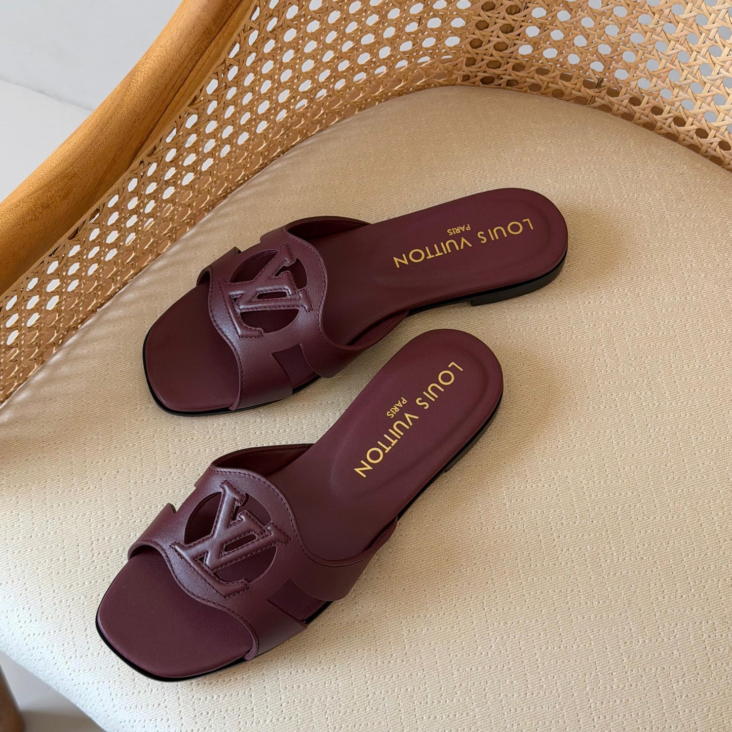 Pair of maroon Louis Vuitton slides on a beige cushioned chair.