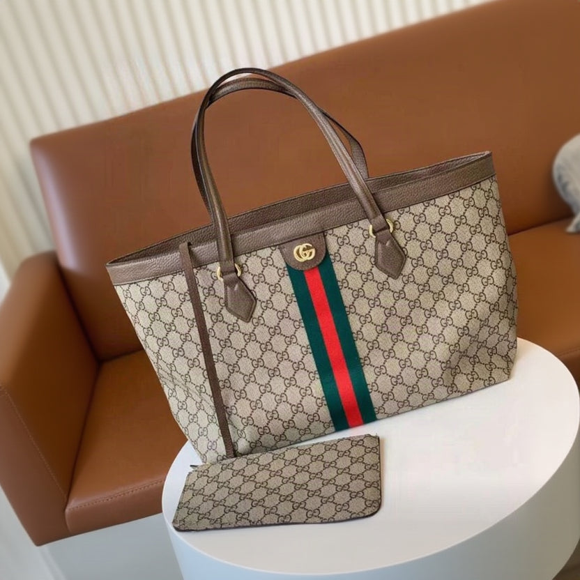 Gucci GG Supreme canvas tote bag with brown leather handles and trim, featuring the signature green and red web stripe and gold interlocking G logo, shown with a matching pouch on a white table against a brown leather background.